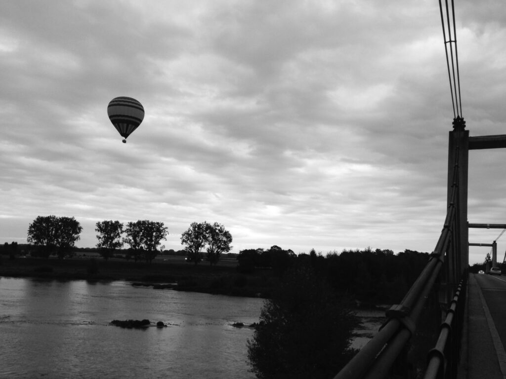 A hot-air balloon in a cloudy sky above a river. The photo was taken from a suspension bridge, a portion of which is visible on the right side of the image.